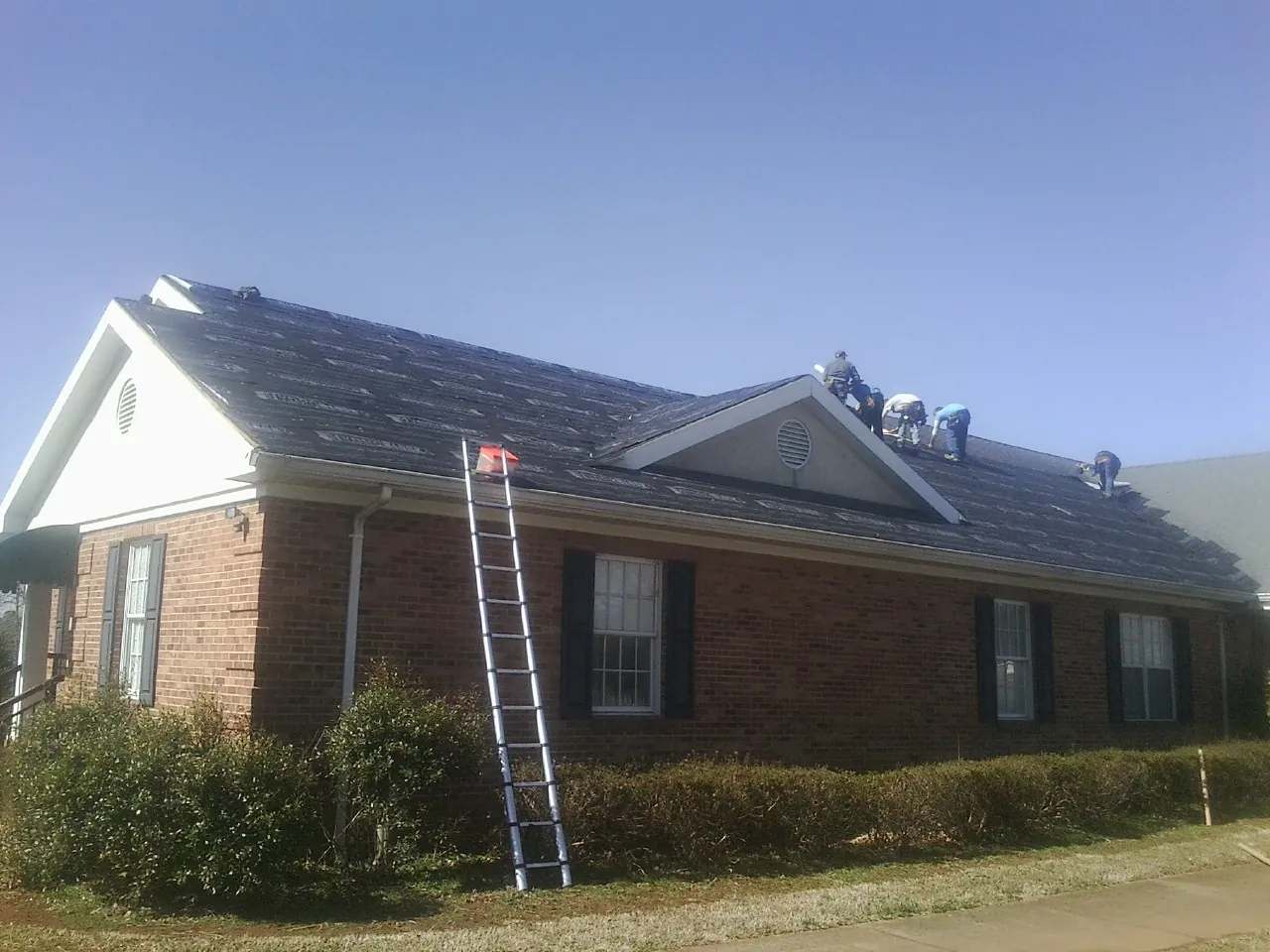 Crew tearing shingles off a brick ranch roof with a ladder set — Metal Roof Repair work by our crew in Honeoye