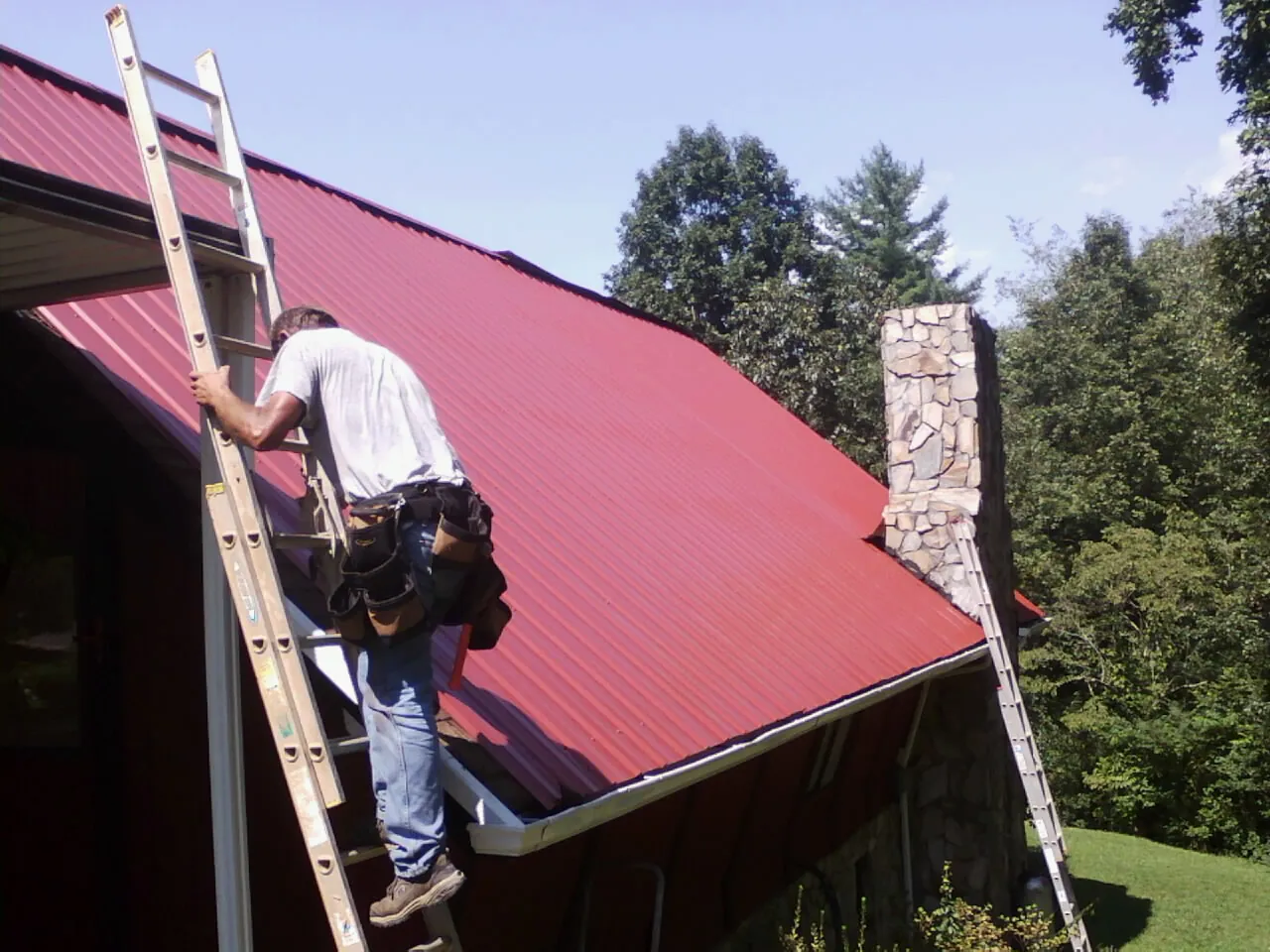 Roofer on a ladder beside a red metal roof and stone chimney — Soffit & Fascia Repair by Roofing Brothers in Honeoye