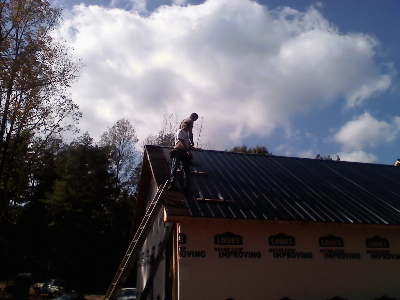 Roofer fastening dark metal panels high against a cloudy sky, from a Roof Inspection job in Honeoye